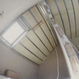 Skośne poddasze z oknem dachowym w trakcie izolacji wełną mineralną, widoczne metalowe profile konstrukcyjne i fragmenty muru z resztkami tynku.