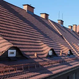 Czerwono-brązowy dach z dachówką karpiówką na budynku z oknami dachowymi i ceglanymi kominami na tle błękitnego nieba.