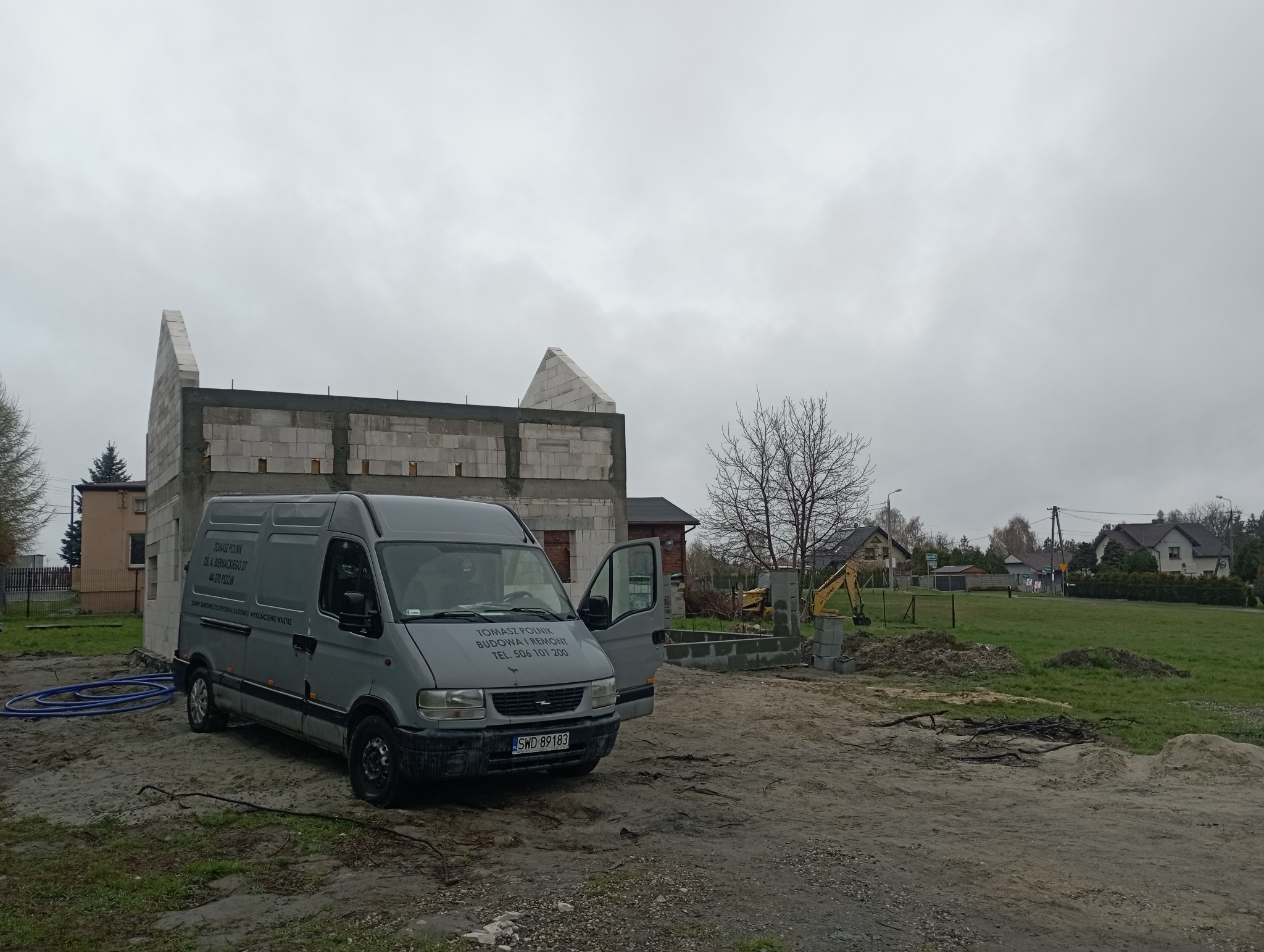 Szara furgonetka firmy budowlanej na placu budowy, widoczny surowy stan budynku z pustaków, częściowo wzniesione ściany, otwarte drzwi boczne furgonetki z widocznym logo firmy i numerem telefonu.