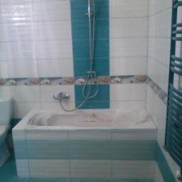 firma jednoosobowa - Wykończona łazienka z wanną obudowaną płytkami w odcieniach bieli i turkusu, kabina prysznicowa z deszczownicą, dekoracyjny pas płytek z motywem muszelek, grzejnik drabinkowy.