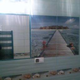 firma jednoosobowa - Wykończenie ściany łazienki z motywem morskiego krajobrazu na płytkach, dekoracyjny pas z motywem muszli, grzejnik drabinkowy po lewej.