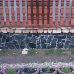 Kamienna elewacja domu z ciemnych, nieregularnych płyt, jasny pas kamienia pod drewnianą balustradą, rabaty kwiatowe z przodu.