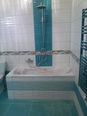 Wykończona łazienka z wanną obudowaną płytkami w odcieniach bieli i turkusu, kabina prysznicowa z deszczownicą, dekoracyjny pas płytek z motywem muszelek, grzejnik drabinkowy.