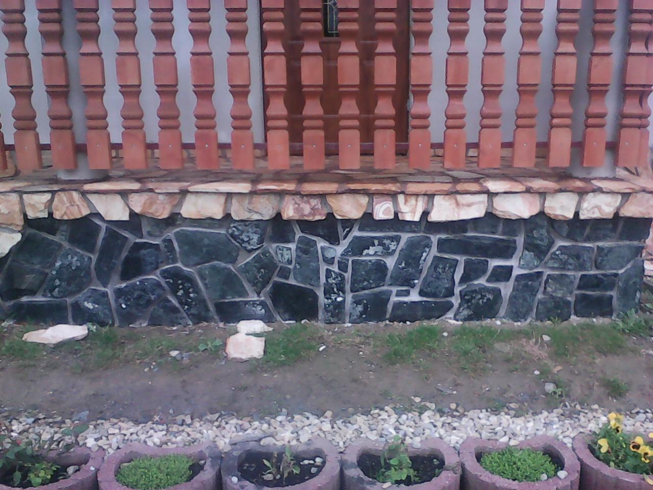 Kamienna elewacja domu z ciemnych, nieregularnych płyt, jasny pas kamienia pod drewnianą balustradą, rabaty kwiatowe z przodu.