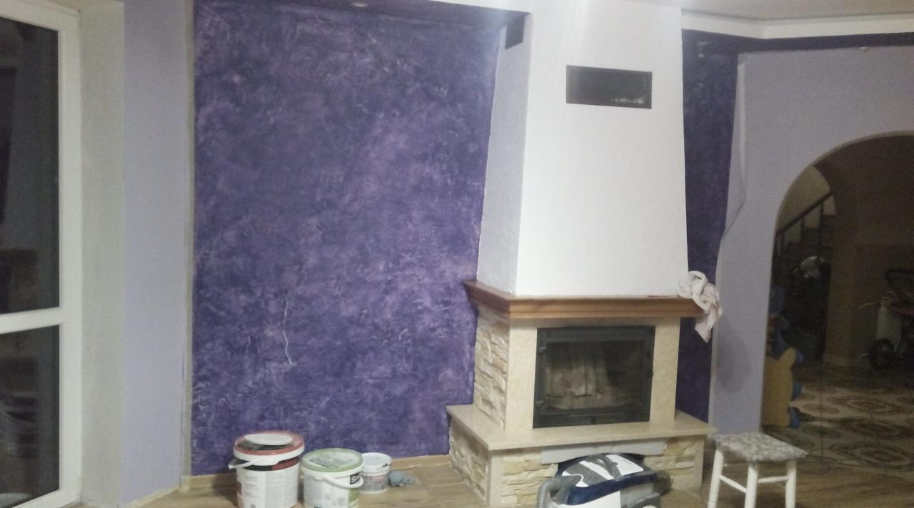 Ściana z fioletową, dekoracyjną strukturą obok kominka z kamienia i drewna. Widoczne puszki z farbą i narzędzia malarskie.