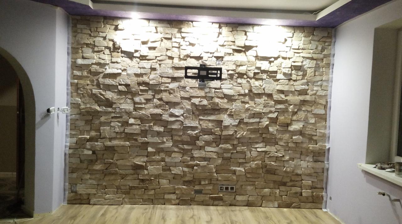 Ściana wyłożona dekoracyjnym kamieniem w salonie, z zamontowanym uchwytem na telewizor i widocznymi gniazdkami elektrycznymi u dołu, oświetlona od góry.