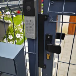 Szary wideofon Vidos z klawiaturą numeryczną zamontowany na metalowej bramie posesji z czarną klamką i zamkiem, widoczna skrzynka na listy i fragment chodnika.