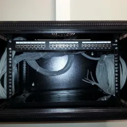 Czarna szafa rack z panelem krosowniczym Cat.5e i uporządkowanymi kablami sieciowymi, widoczne numery portów 1-24.