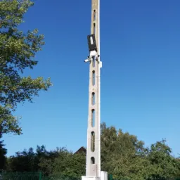 Betonowy słup z zamontowanymi kamerami monitoringu i oświetleniem, widoczna skrzynka elektryczna u podstawy, na tle błękitnego nieba i zieleni.