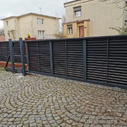 Nowoczesna, przesuwna brama z poziomymi lamelami w kolorze antracytowym, zamontowana na posesji z brukowaną nawierzchnią i ceglanym ogrodzeniem, widoczny napęd elektryczny bramy.