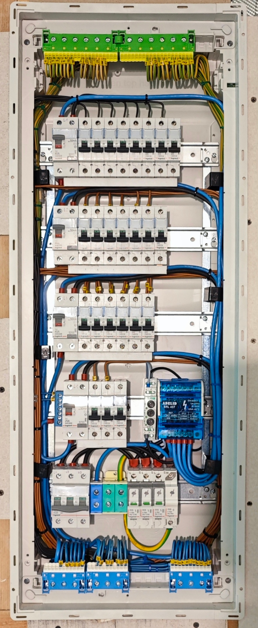 Wnętrze rozdzielnicy elektrycznej z precyzyjnie ułożonymi przewodami w kolorach niebieskim, brązowym i żółto-zielonym, widoczne bezpieczniki i moduł przepięciowy Adelid BRL 407, całość w białej...
