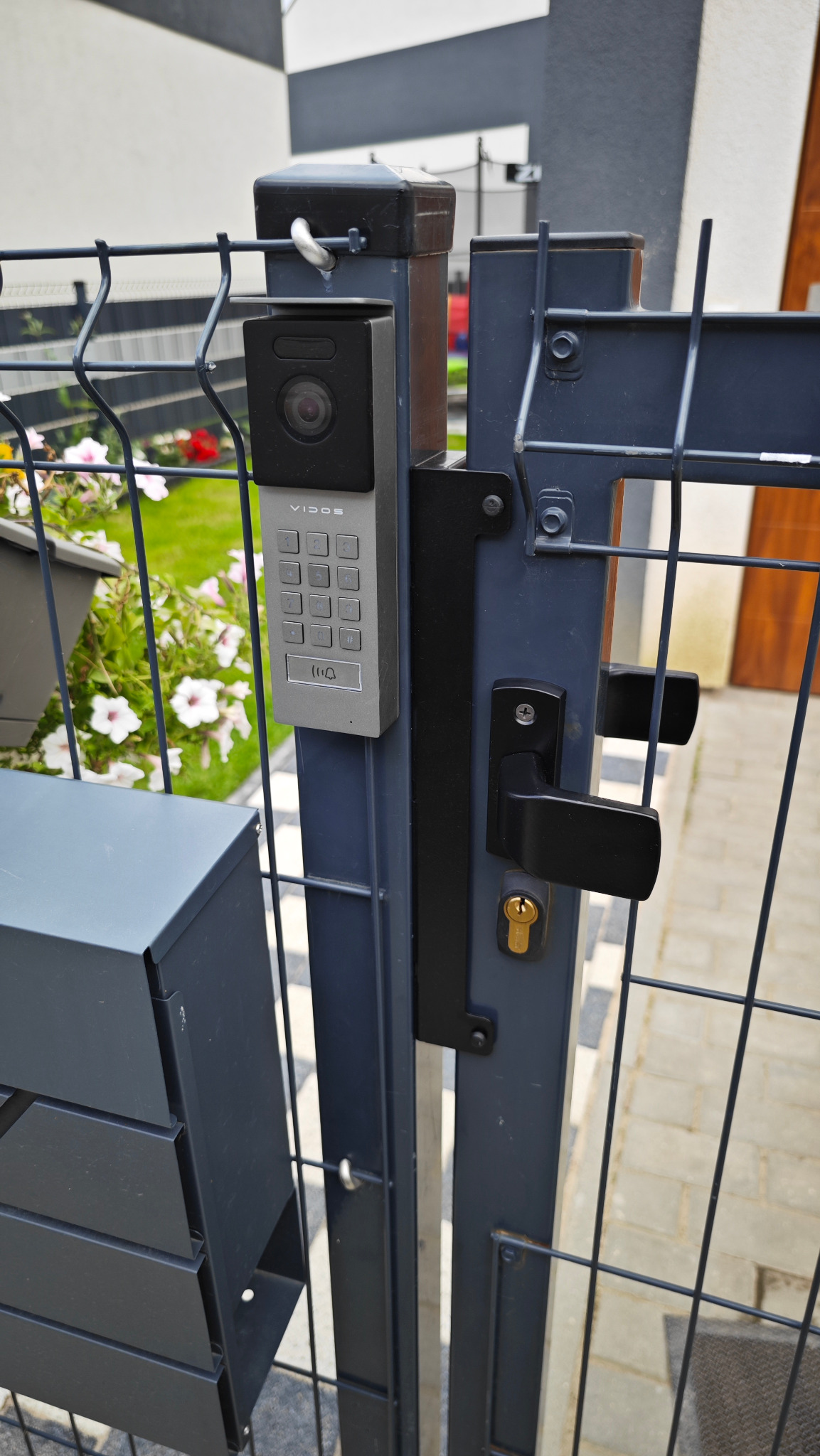Szary wideofon Vidos z klawiaturą numeryczną zamontowany na metalowej bramie posesji z czarną klamką i zamkiem, widoczna skrzynka na listy i fragment chodnika.