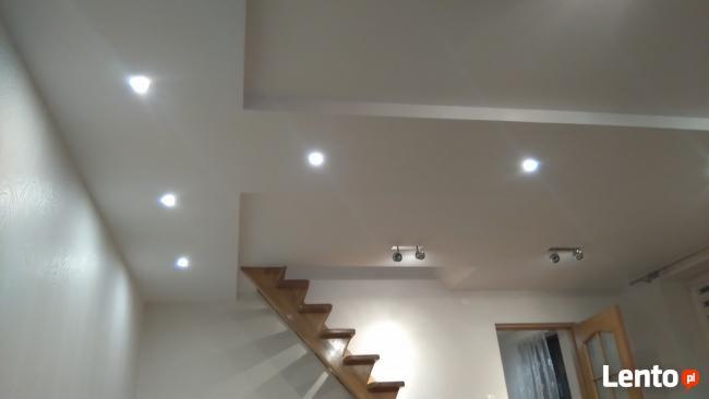 Biały sufit z wbudowanymi punktowymi lampami LED, drewniane schody w tle, widok na świeżo odnowione wnętrze.