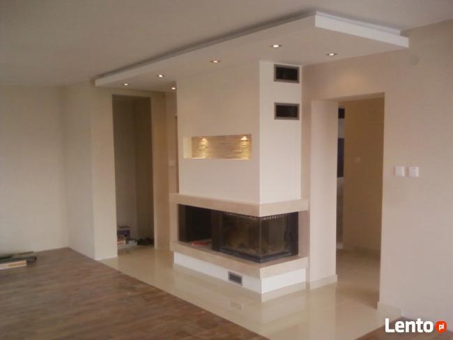 Wykończone wnętrze z nowoczesnym kominkiem obudowanym jasnym kamieniem, widoczne elementy sufitu podwieszanego z oświetleniem punktowym i świeżo pomalowane ściany w odcieniach beżu.