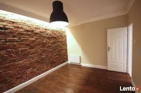 Pusty pokój z odnowioną ceglaną ścianą, błyszczącą drewnianą podłogą, białymi listwami przypodłogowymi, białymi drzwiami i wiszącą czarną lampą.