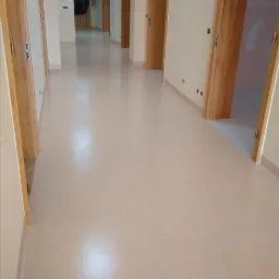 Długi korytarz z beżową, jednolitą podłogą, widoczne drewniane ościeżnice drzwi po obu stronach, perspektywa zbieżna w głąb.