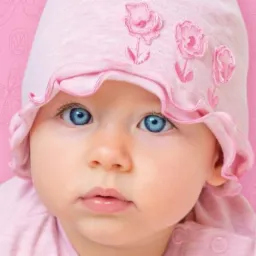 Portret niemowlęcia z intensywnie niebieskimi oczami w różowej czapeczce z aplikacjami kwiatowymi na tle z motywem motyli i kwiatów.
