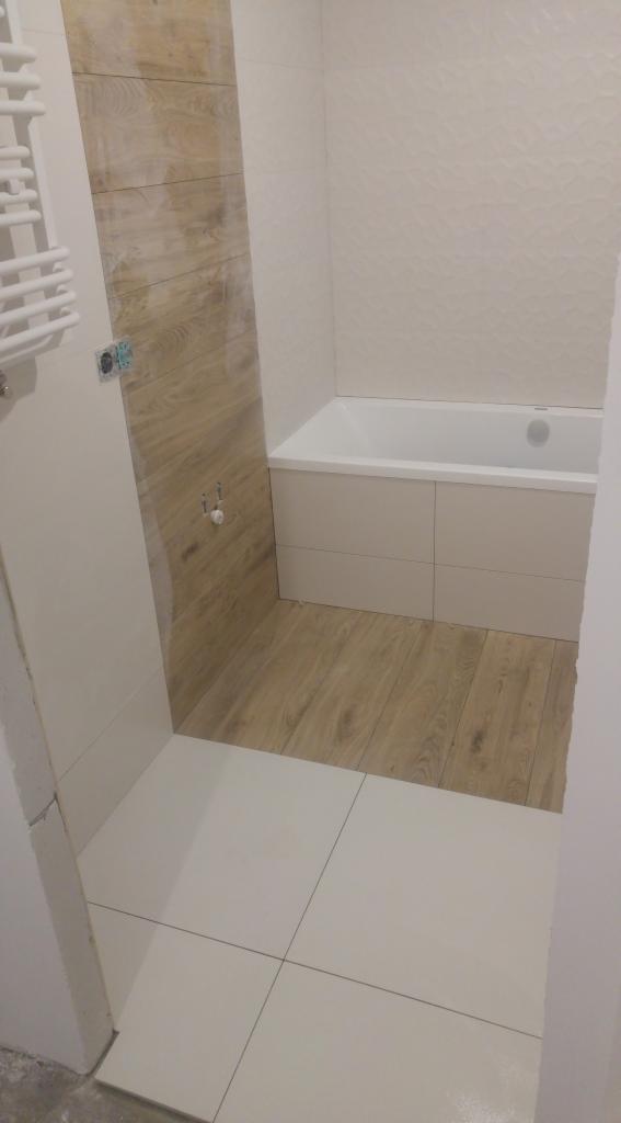 Wykończona łazienka z drewnopodobnymi płytkami na ścianie i podłodze, biała wanna obudowana płytkami, grzejnik drabinkowy po lewej stronie.