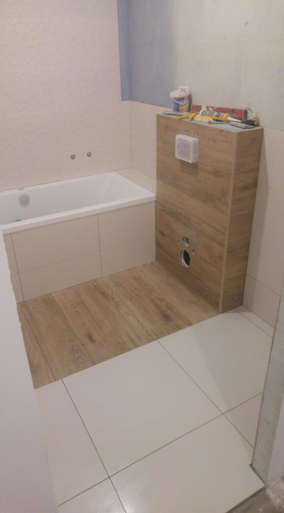 Niedokończona łazienka z wanną w zabudowie z białych płytek, podłogą z paneli imitujących drewno i częściowo wyłożonymi białymi płytkami, widoczna zabudowa stelaża WC z drewnopodobnych paneli,...