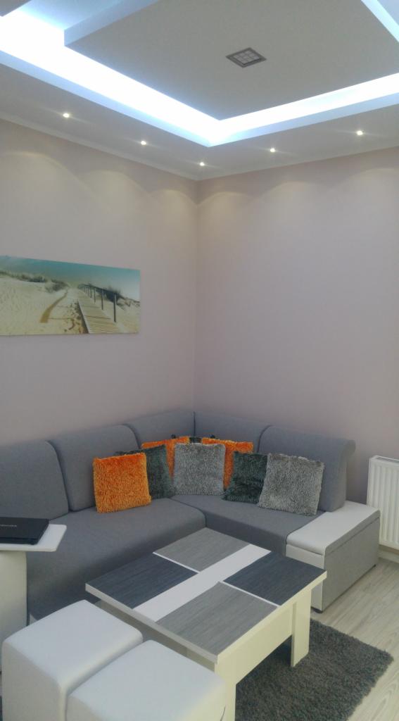 Jasne wnętrze salonu z narożną sofą, stolikiem kawowym z szarymi akcentami i dekoracyjnym oświetleniem sufitowym.