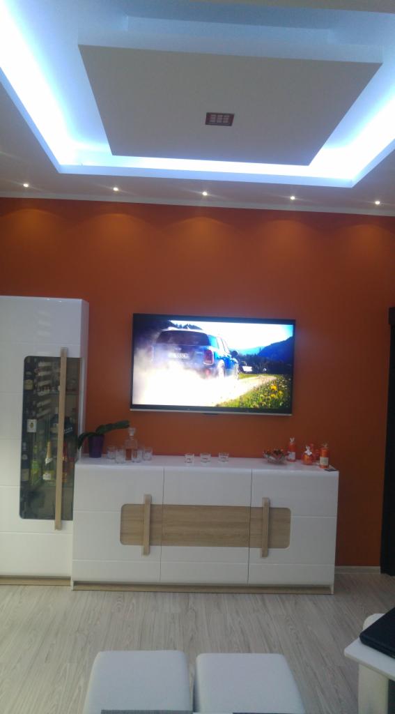 Pomarańczowa ściana w salonie z białym kredensem, telewizorem z widokiem na krajobraz i podwieszanym sufitem z oświetleniem LED.