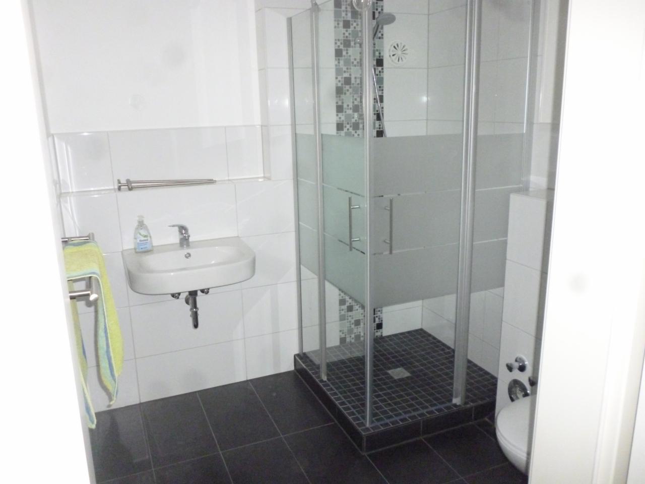 Wykończona łazienka z kabiną prysznicową z częściowo matowym szkłem, umywalką wiszącą i czarną podłogą z płytek. Na ścianach białe płytki, a nad kabiną prysznicową dekoracyjny pas z mozaiki.