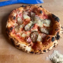 Okrągła pizza z lekko przypalonym brzegiem, z szynką, serem i plasterkami ogórka konserwowego, ułożona na drewnianej desce obok rozsypanej mąki. W tle niebieska łopatka do pizzy.