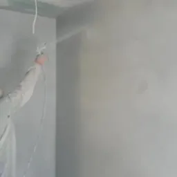Pracownik w kombinezonie ochronnym nanosi farbę metodą natryskową na ścianę wewnątrz pomieszczenia, widoczne ślady świeżo nałożonej powłoki i przewód zasilający pistolet natryskowy.