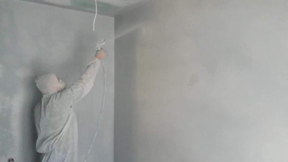 Pracownik w kombinezonie ochronnym nanosi farbę metodą natryskową na ścianę wewnątrz pomieszczenia, widoczne ślady świeżo nałożonej powłoki i przewód zasilający pistolet natryskowy.