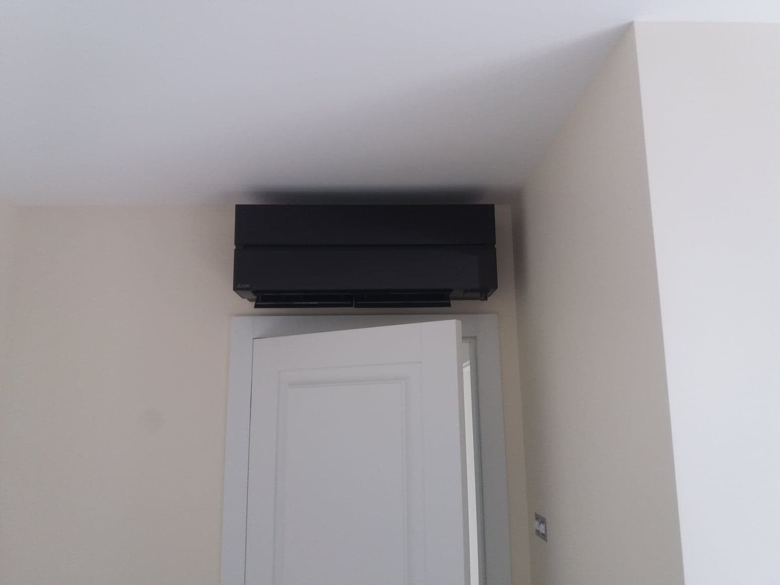 Czarna, pozioma jednostka klimatyzacji Mitsubishi zamontowana nad otwartymi, białymi drzwiami w jasnym pomieszczeniu z kremowymi ścianami.