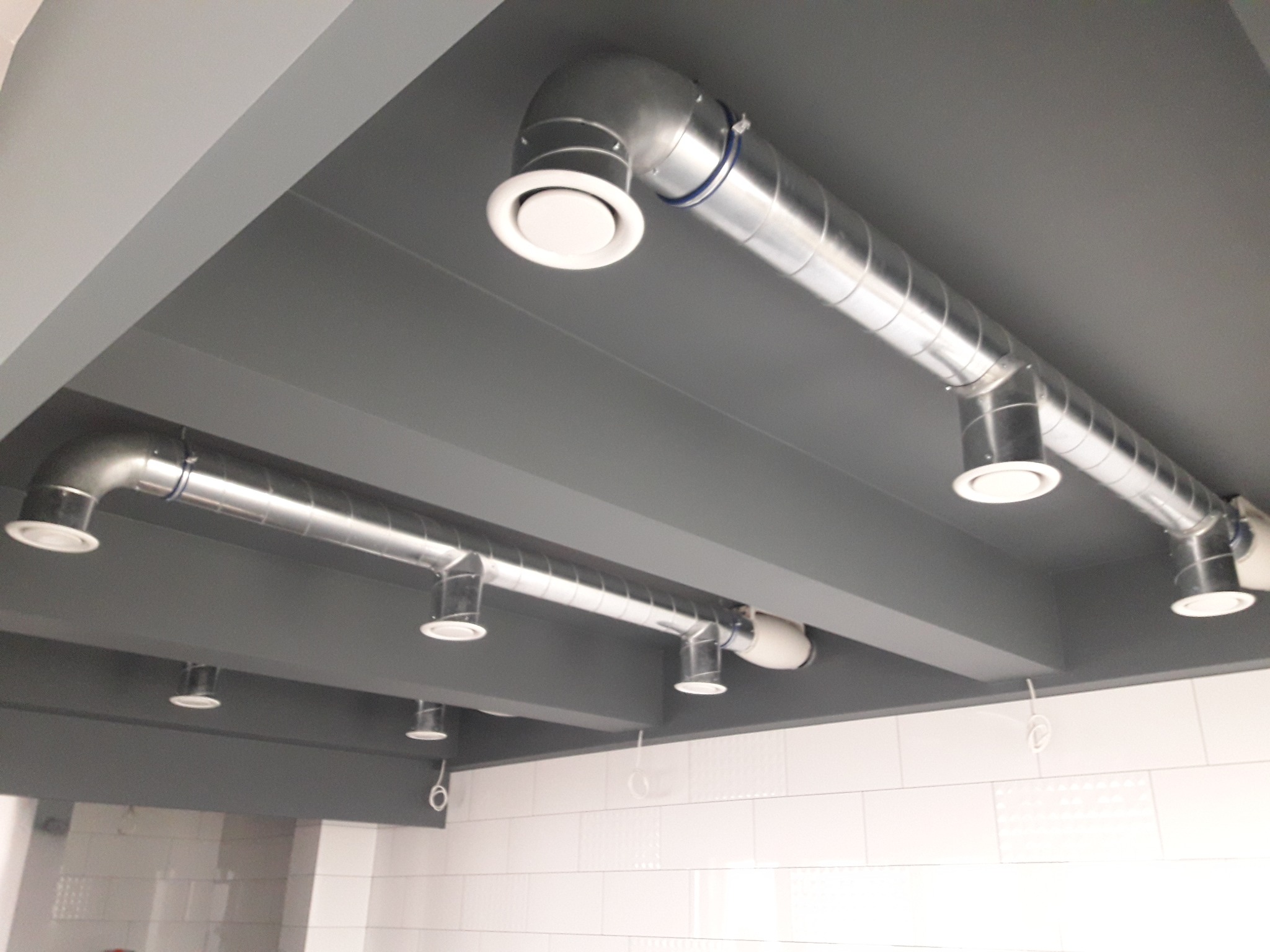Szare pomieszczenie z widoczną instalacją wentylacyjną pod sufitem, składającą się z metalowych rur i białych okrągłych nawiewników.