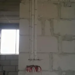 Białe przewody elektryczne przymocowane klamrami do ściany z betonu komórkowego, widoczne wystające kable ze ściany w miejscu gniazdka, okno w tle.