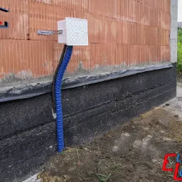 Zewnętrzna skrzynka elektryczna z niebieską rurą karbowaną przymocowana do ściany budynku z cegły, widoczne czarne zabezpieczenie fundamentów.