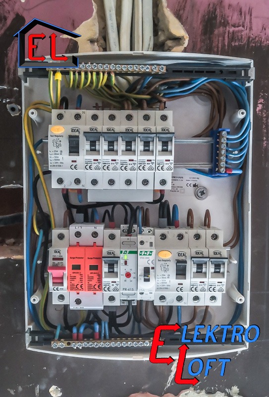 Otwarta skrzynka rozdzielcza z bezpiecznikami i przewodami w różnych kolorach, widoczne zabezpieczenia przeciwprzepięciowe i listwy zaciskowe, zamontowana na nierównej ścianie z widocznymi śladami...