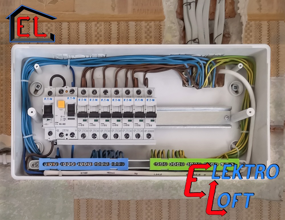 Wnętrze białej skrzynki rozdzielczej z zainstalowanymi bezpiecznikami EATON, widoczne przewody w różnych kolorach (niebieski, brązowy, zielono-żółty), listwy zaciskowe w kolorze niebieskim...