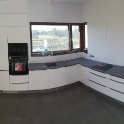 Nowoczesna, biała kuchnia z szarym blatem, widok na okno z zielenią za nim, częściowo wykończona, z widocznymi niedokończonymi elementami instalacji elektrycznej.