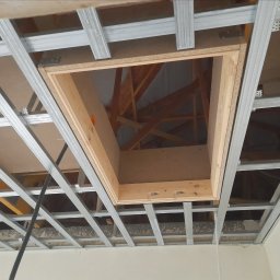 remonty wykończenia wnetrz - Stelaż sufitowy z zamontowanym drewnianym otworem rewizyjnym, widoczne elementy konstrukcji dachu.
