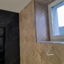 Ściana wyłożona drewnopodobnymi płytkami ułożonymi w jodełkę, obok ściana z czarnego marmuru z białymi żyłami, widoczne okno i wystający kabel.