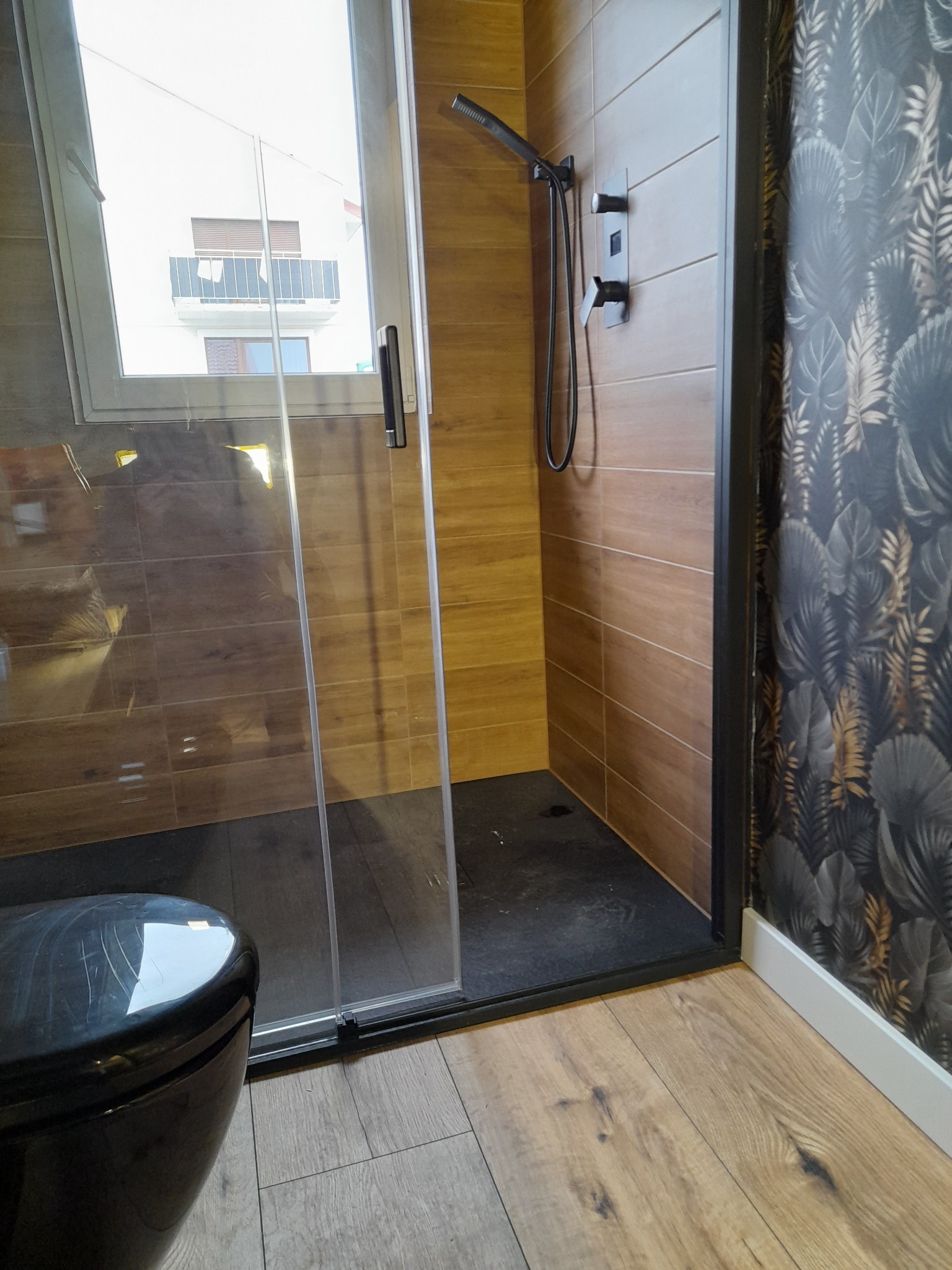 Nowoczesna kabina prysznicowa z drewnopodobnymi płytkami i czarną armaturą, widok na zewnątrz przez okno w kabinie, wzorzysta tapeta w tle.