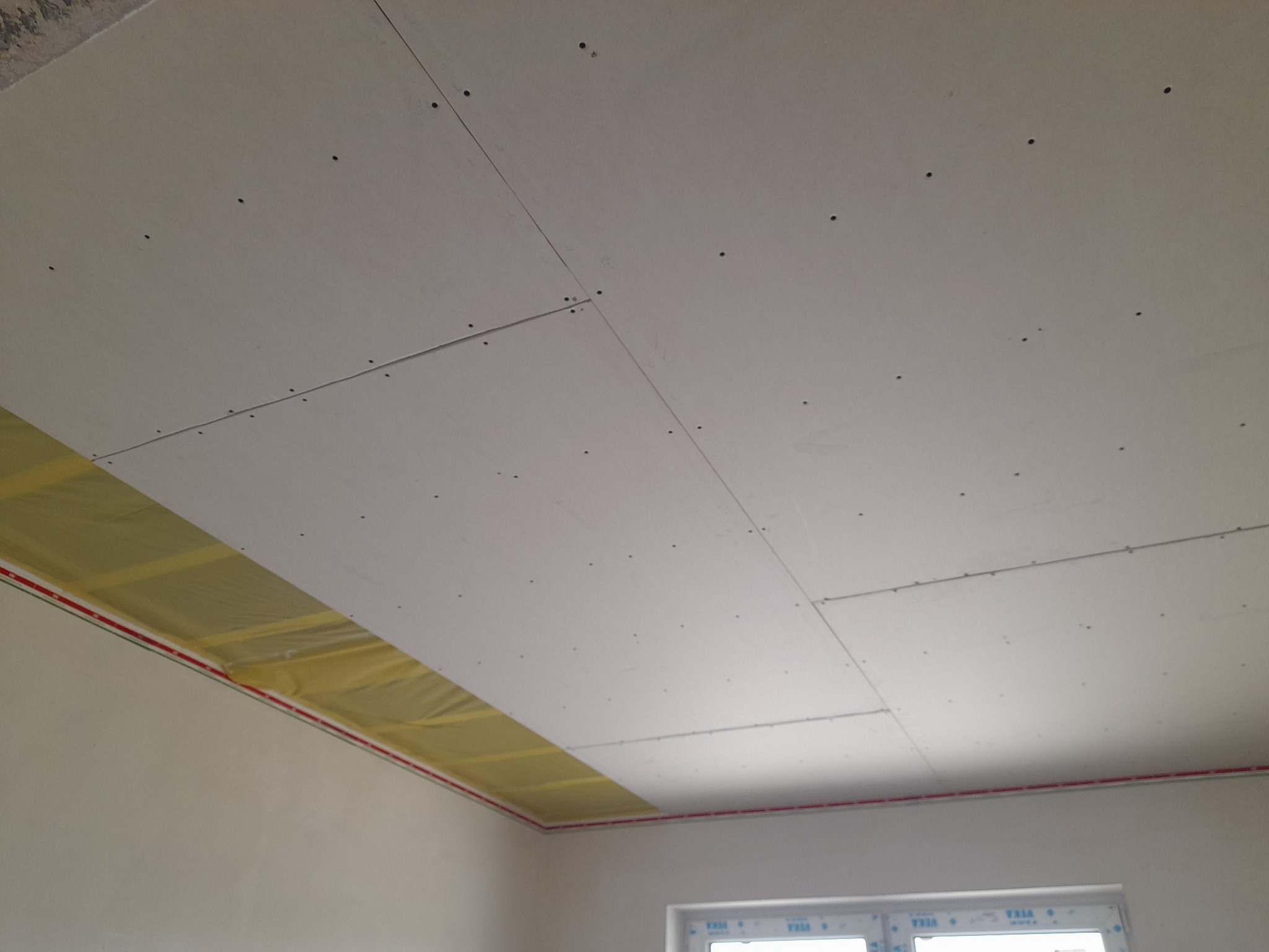 Sufit podwieszany z płyt gipsowo-kartonowych, widoczne łączenia płyt i wkręty, częściowo ocieplony wełną mineralną z żółtą folią, czerwona taśma oddzielająca sufit od ściany.