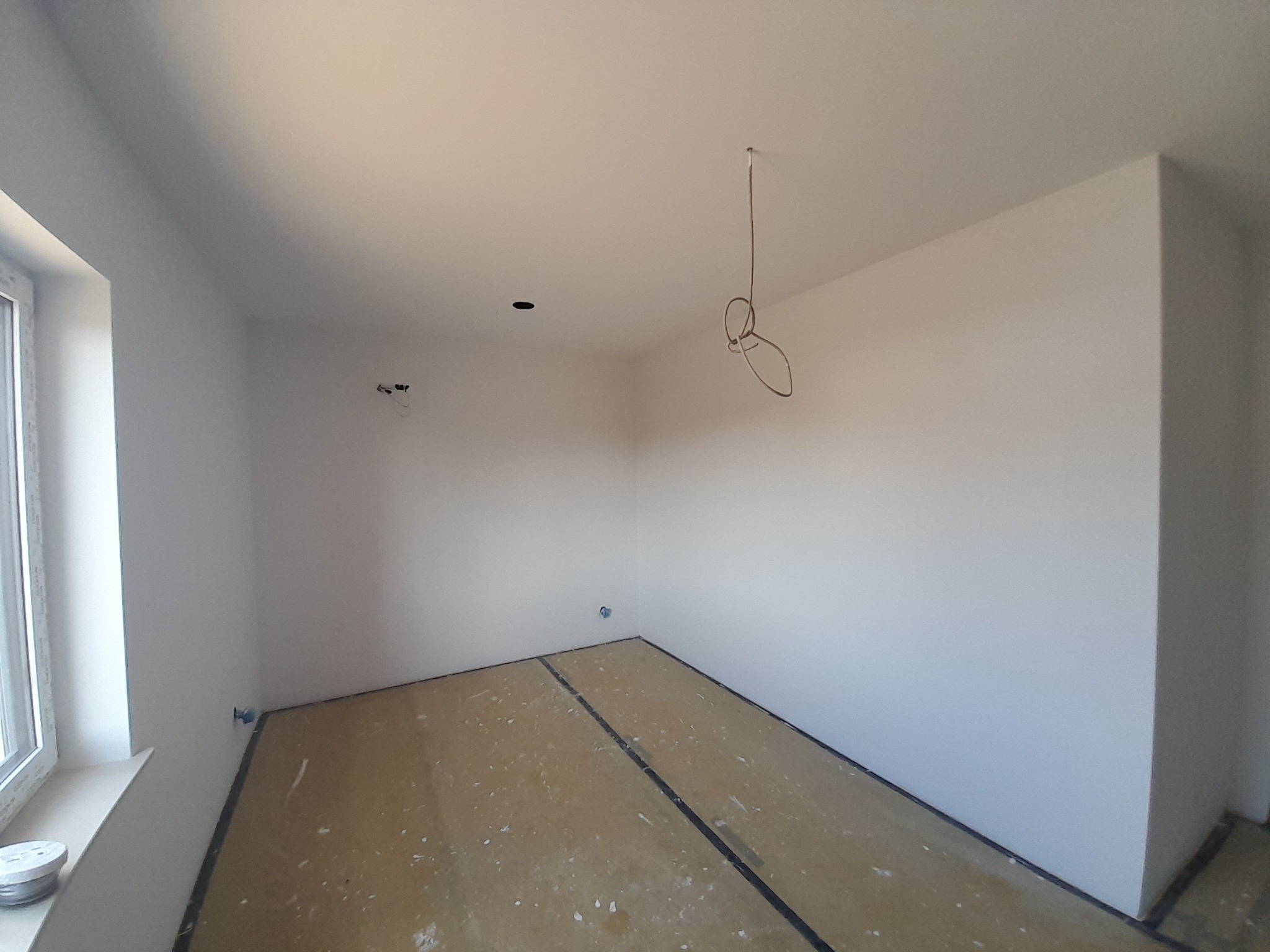 Pomalowane na biało wnętrze pokoju w trakcie prac wykończeniowych, zabezpieczona podłoga, widoczne wystające przewody elektryczne, okno po lewej stronie.