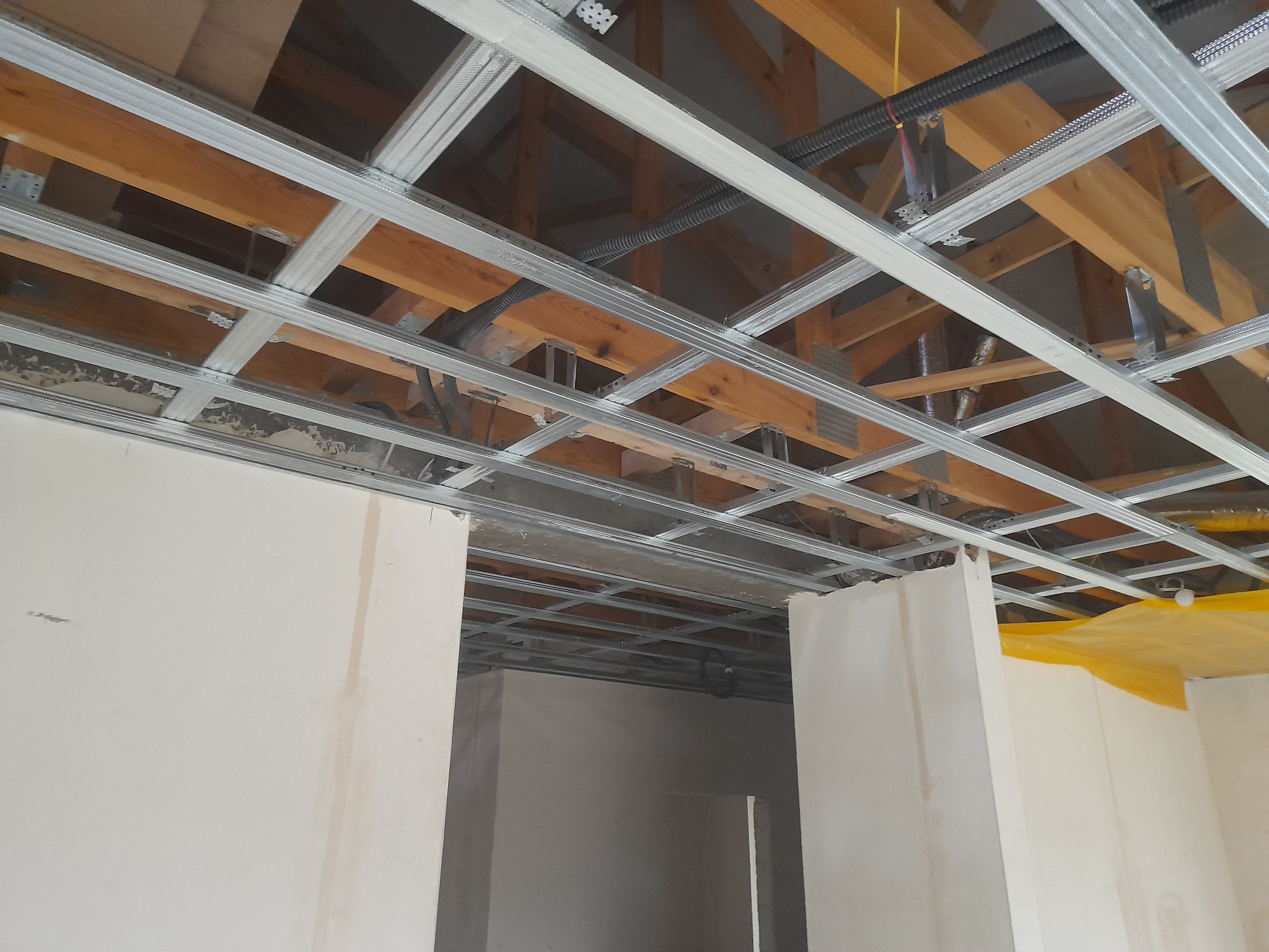 Konstrukcja metalowa podwieszanego sufitu zamontowana do drewnianej więźby dachowej, widoczne przewody elektryczne i fragmenty ścian w stanie surowym.