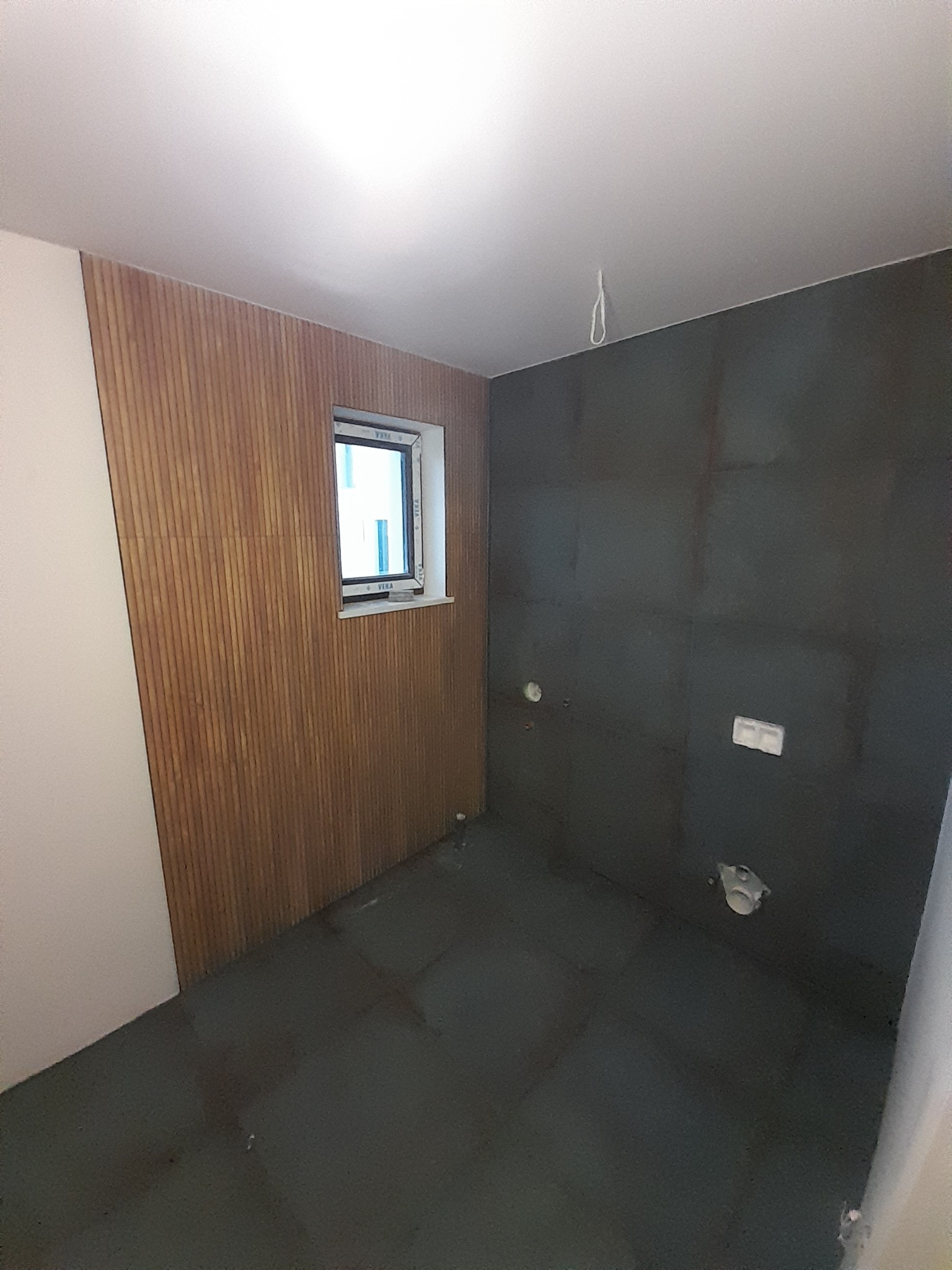 Łazienka w trakcie prac wykończeniowych: ściana z drewnianymi panelami pionowymi, okno, surowa ściana w ciemnym kolorze z widocznymi instalacjami, podłoga pokryta folią ochronną.