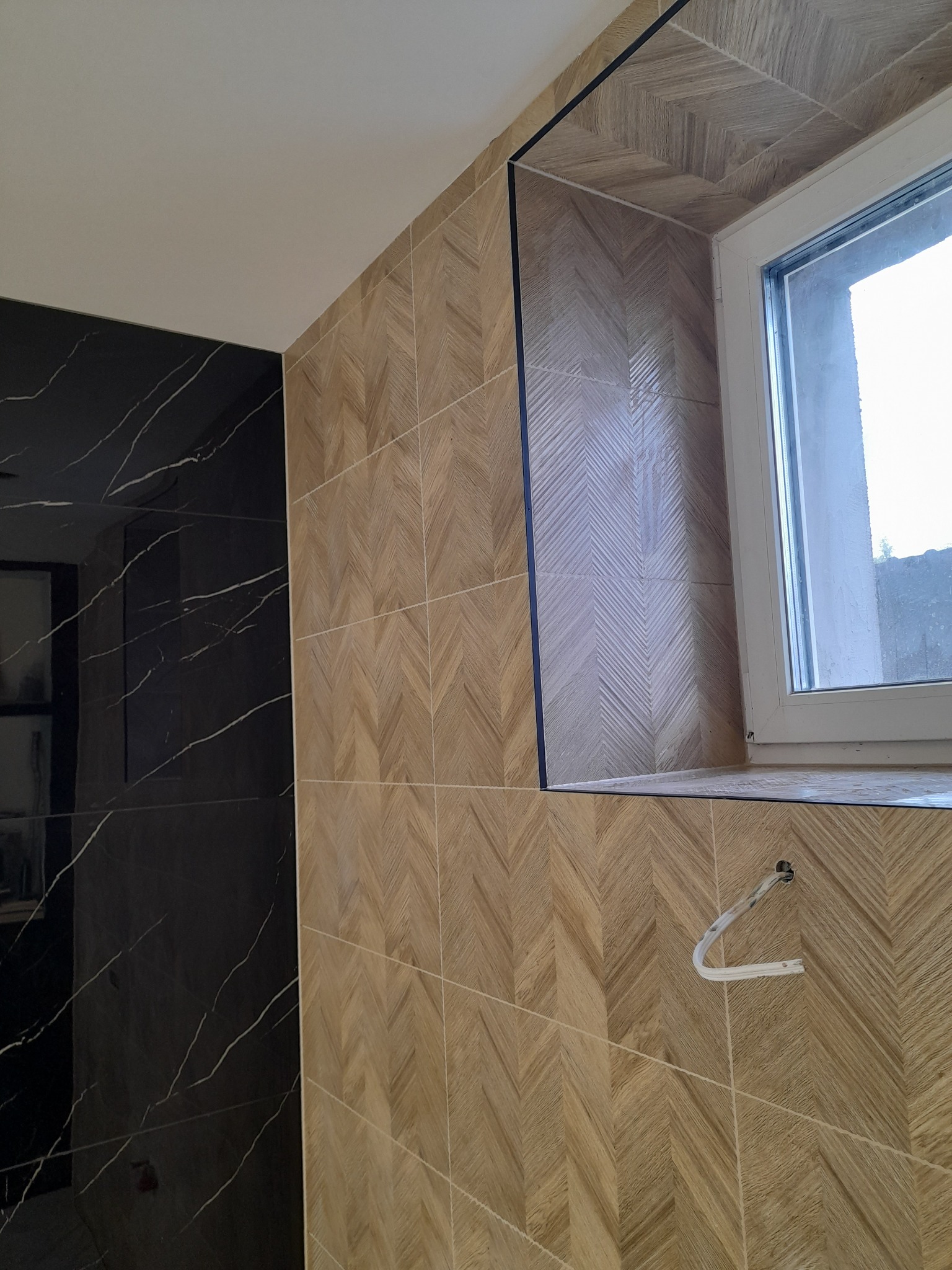Ściana wyłożona drewnopodobnymi płytkami ułożonymi w jodełkę, obok ściana z czarnego marmuru z białymi żyłami, widoczne okno i wystający kabel.