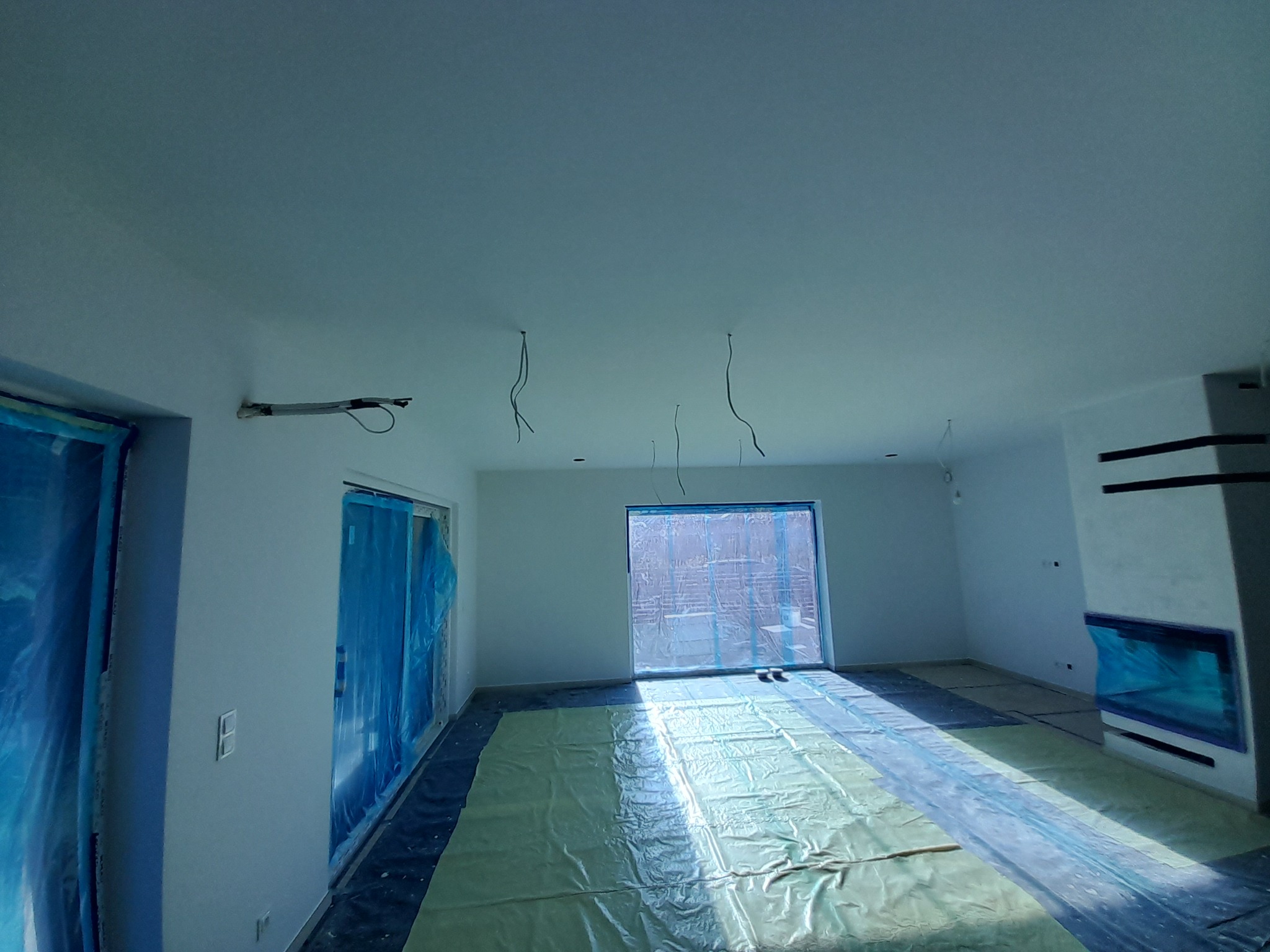 Białe wnętrze po świeżym malowaniu, zabezpieczone folią. Widoczne wiszące kable, okna i drzwi osłonięte folią malarską. Minimalistyczny kominek z prawej strony.