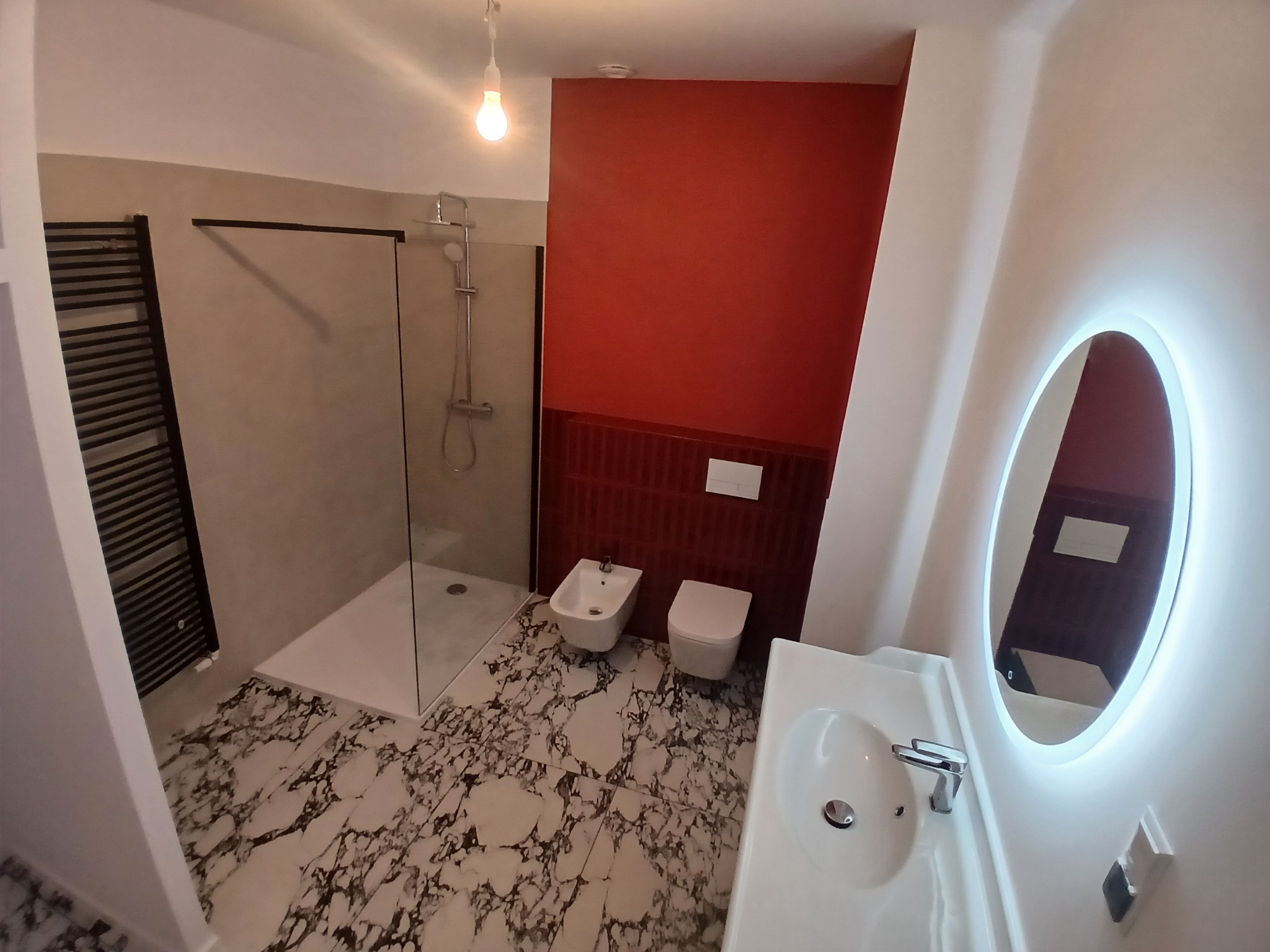 Nowoczesna łazienka z kabiną prysznicową, bidetem, toaletą i umywalką. Czerwona ściana z dekoracyjnymi płytkami. Podłoga z marmurowym wzorem. Lustro z podświetleniem LED.