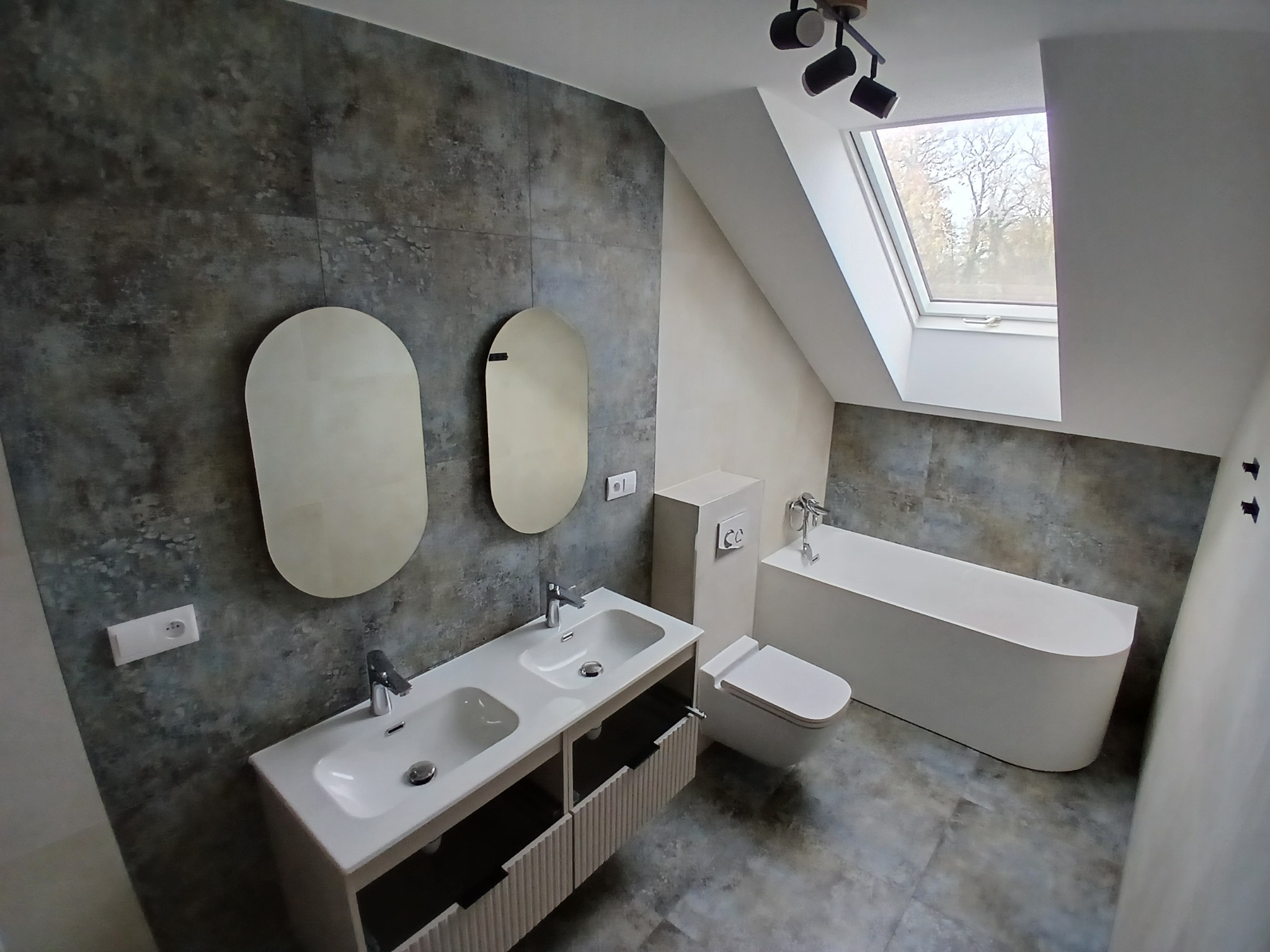 Nowoczesna łazienka na poddaszu z dwoma umywalkami, owalnymi lustrami i wanną wolnostojącą. Szare płytki imitujące kamień na ścianach i podłodze.