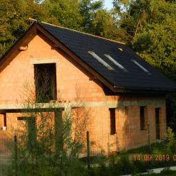 Michalski - Niedokończony dom z czerwonej cegły z nowym, czarnym dachem z trzema oknami dachowymi, widoczny przez ogrodzenie i roślinność, z datą wykonania zdjęcia w prawym dolnym rogu.