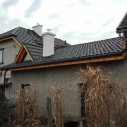 Dom z nowym, grafitowym dachem z dachówki ceramicznej, wykończonym drewnianą podbitką, widoczne białe kominy i rynny, na pierwszym planie wysokie, brązowe trawy.