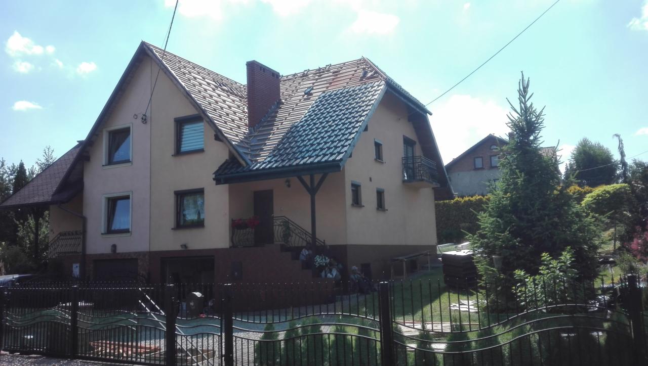 Dwupiętrowy dom z częściowo zdjętym dachem, widoczny fragment nowej dachówki i odsłonięta konstrukcja, na pierwszym planie ogrodzenie posesji.